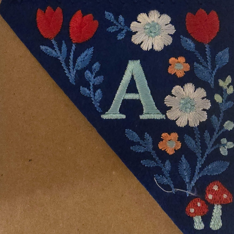 May include: A blue felt banner with embroidered floral and mushroom designs. The banner features the letter "A" in light blue, surrounded by red tulips, white and orange flowers, and blue leaves. Two red mushrooms with white spots are at the bottom.