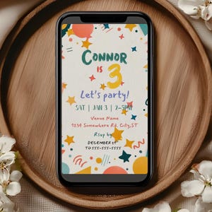 Op de afbeelding: Een smartphone toont een kleurrijke uitnodiging voor een verjaardagsfeestje. De uitnodiging luidt "Connor is 3" met de tekst "Laten we feesten!" en evenementdetails. De telefoon ligt op een houten dienblad, omringd door witte bloemen.