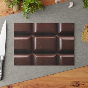 May include: A rectangular chopping board with a close-up of a dark chocolate bar. The chocolate is divided into nine squares, with a glossy, textured surface. The board is on a grey surface with a knife and greenery.