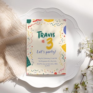 May include: A birthday party invitation with the text "Travis is 3, Let's party!" on a cream-colored background. The invitation is decorated with colorful shapes and placed on a white plate, with a sprig of white flowers.