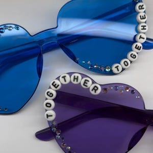May include: Heart-shaped sunglasses in blue and purple. The blue pair has the word "TOGETHER" spelled out in white beads along the frame. Both pairs are embellished with small, sparkling rhinestones.