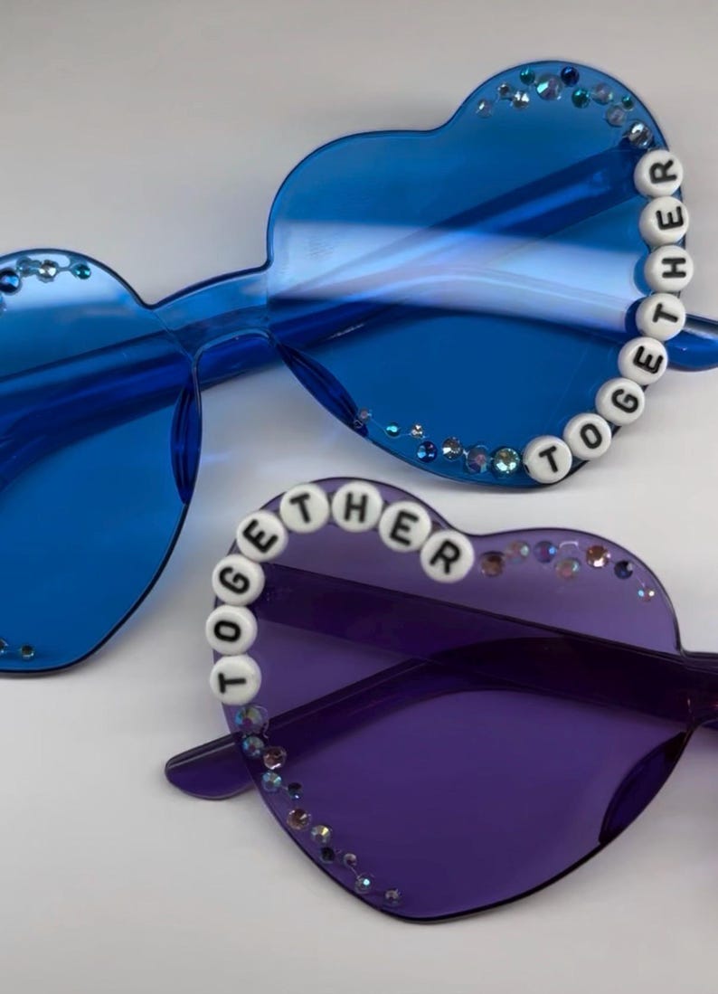May include: Heart-shaped sunglasses in blue and purple. The blue pair has the word "TOGETHER" spelled out in white beads along the frame. Both pairs are embellished with small, sparkling rhinestones.