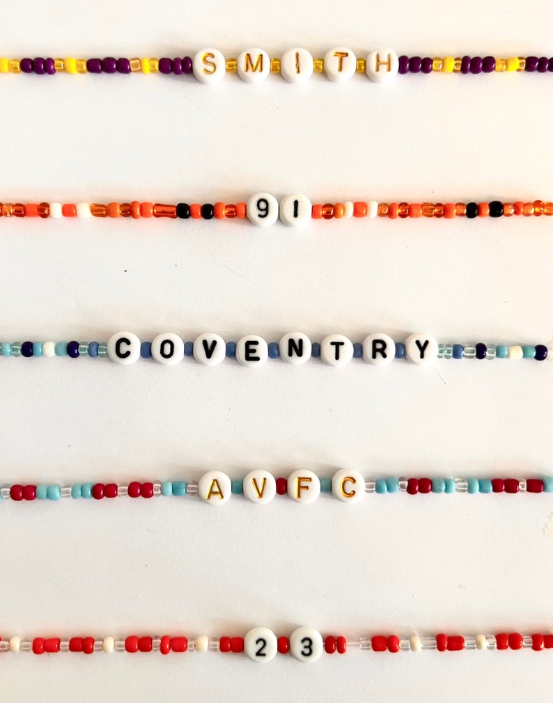 Puede incluir: Una colecci&oacute;n de pulseras de cuentas con cuentas blancas con letras y n&uacute;meros. Las pulseras muestran las palabras "SMITH", "COVENTRY" y "AVFC", junto con los n&uacute;meros "91" y "23". Las cuentas son de varios colores, incluyendo rojo, naranja y azul.