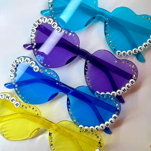 May include: Heart-shaped sunglasses in shades of blue, purple, and yellow. Each pair has the word "TOGETHER" spelled out in white beads along the frame and is embellished with small rhinestones. The lenses are transparent.