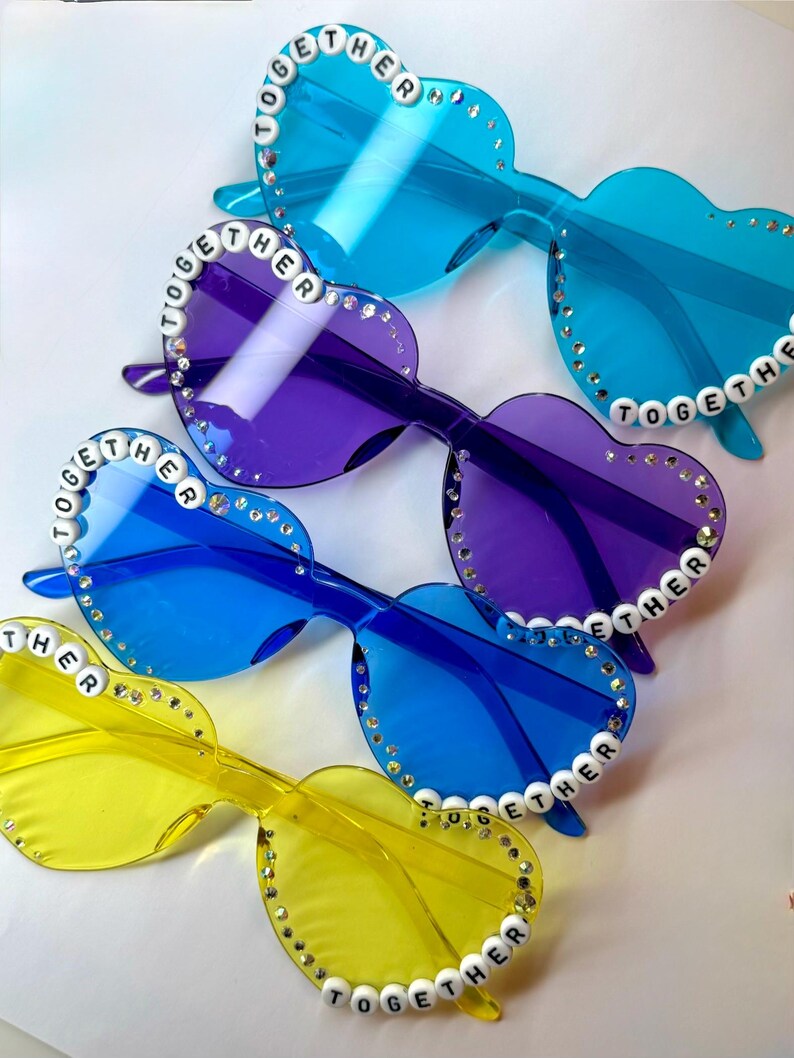 May include: Heart-shaped sunglasses in shades of blue, purple, and yellow. Each pair has the word "TOGETHER" spelled out in white beads along the frame and is embellished with small rhinestones. The lenses are transparent.