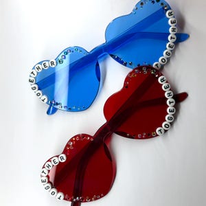 May include: Two pairs of heart-shaped sunglasses. One pair is blue, the other is red. Both have the word "TOGETHER" spelled out in white beads along the frame. The frames are decorated with small, sparkling rhinestones.