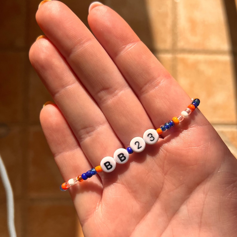 Puede incluir: Una pulsera de cuentas con letras blancas que deletrean "BB23" e intercaladas con cuentas azules, naranjas y transparentes. La pulsera se sostiene en una mano, mostrando el dise&ntilde;o colorido y el peque&ntilde;o tama&ntilde;o de las cuentas.