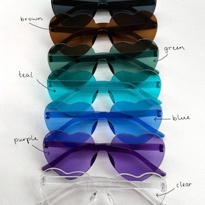 May include: A stack of heart-shaped sunglasses in various colors: black, brown, green, teal, blue, purple, and clear. The sunglasses have a rimless design and are arranged vertically, showcasing the different lens colors.
