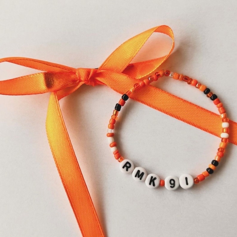 Puede incluir: Una pulsera de cuentas naranjas, negras y blancas, con cuentas de letras blancas que deletrean "RMK91". Una cinta naranja est&aacute; atada en un lazo junto a la pulsera.