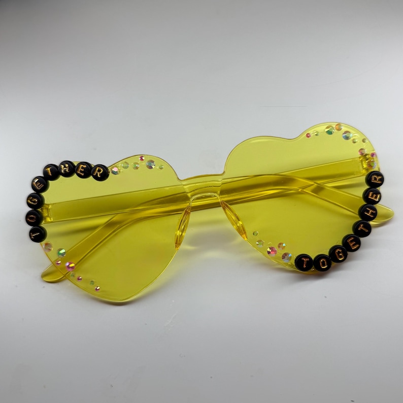May include: Yellow heart-shaped sunglasses with the words "TOGETHER" and "HER" spelled out in black beads. The sunglasses have a clear frame and are decorated with small, iridescent rhinestones.