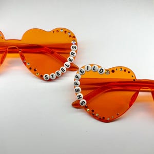 May include: Two pairs of heart-shaped sunglasses in a vibrant orange hue. The frames are adorned with small, sparkling embellishments. One pair spells out "STEELERS" with white letter beads, and the other spells "SHEFFIELD".