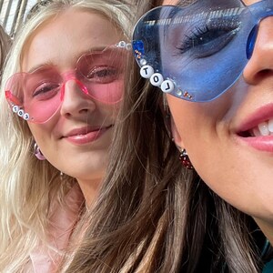 May include: Two people wearing heart-shaped sunglasses. One pair is pink, the other is blue. The blue sunglasses have the letters "TOJ" on the frame. Both people are smiling. The background is a blurred outdoor setting.