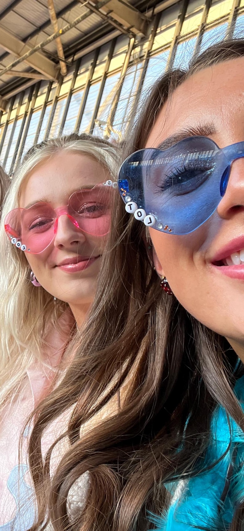 May include: Two people wearing heart-shaped sunglasses. One pair is pink, the other is blue. The blue sunglasses have the letters "TOJ" on the frame. Both people are smiling. The background is a blurred outdoor setting.