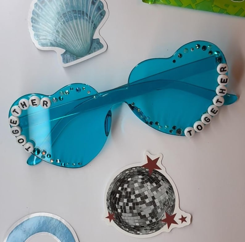 May include: Blue heart-shaped sunglasses with the word "TOGETHER" spelled out in white beads. The sunglasses have a clear frame and are decorated with small rhinestones. A disco ball sticker and a seashell sticker are also visible.