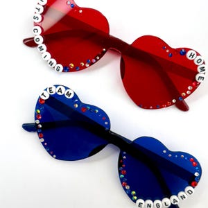 May include: Two pairs of heart-shaped sunglasses. The top pair is red with white beads spelling IT'S COMING HOME and small multicolored rhinestones. The bottom pair is blue with white beads spelling TEAM ENGLAND and small multicolored rhinestones.