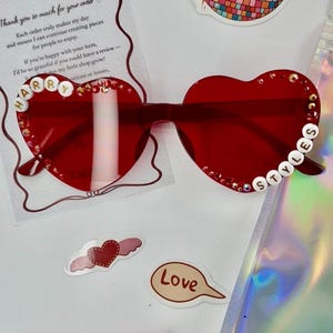 May include: Red heart-shaped sunglasses with the name "HARRY STYLES" spelled out in white beads. The image also includes stickers, a candy bar, and a thank you note.