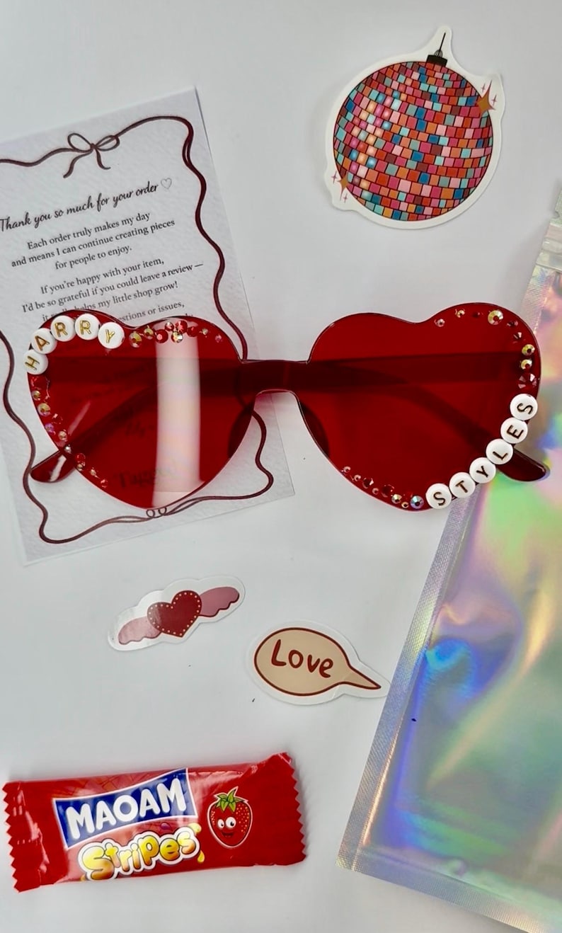 May include: Red heart-shaped sunglasses with the name "HARRY STYLES" spelled out in white beads. The image also includes stickers, a candy bar, and a thank you note.