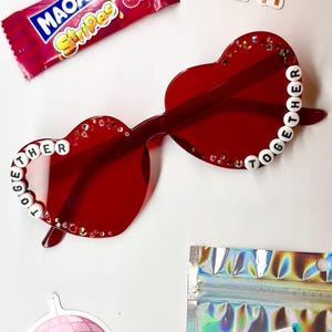 May include: Red heart-shaped sunglasses with the word "TOGETHER" spelled out in white beads. The sunglasses are decorated with small rhinestones. A candy wrapper, stickers, and a disco ball sticker are also visible.