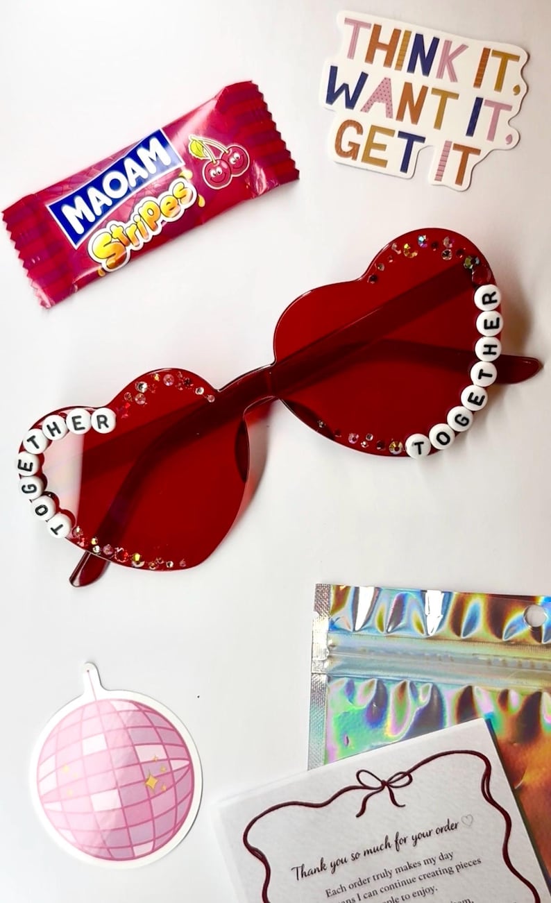 May include: Red heart-shaped sunglasses with the word "TOGETHER" spelled out in white beads. The sunglasses are decorated with small rhinestones. A candy wrapper, stickers, and a disco ball sticker are also visible.
