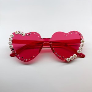 May include: Bright red heart-shaped sunglasses with the phrase "KISS ALL THE TIME" spelled out in white letter beads. The frames are adorned with small, sparkling rhinestones.