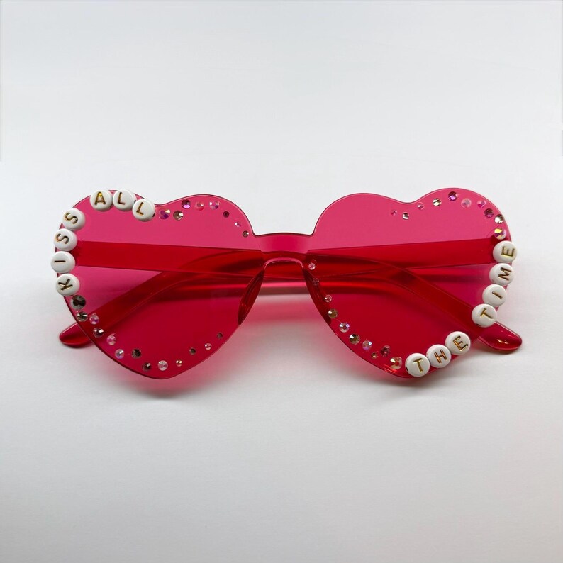 May include: Bright red heart-shaped sunglasses with the phrase "KISS ALL THE TIME" spelled out in white letter beads. The frames are adorned with small, sparkling rhinestones.