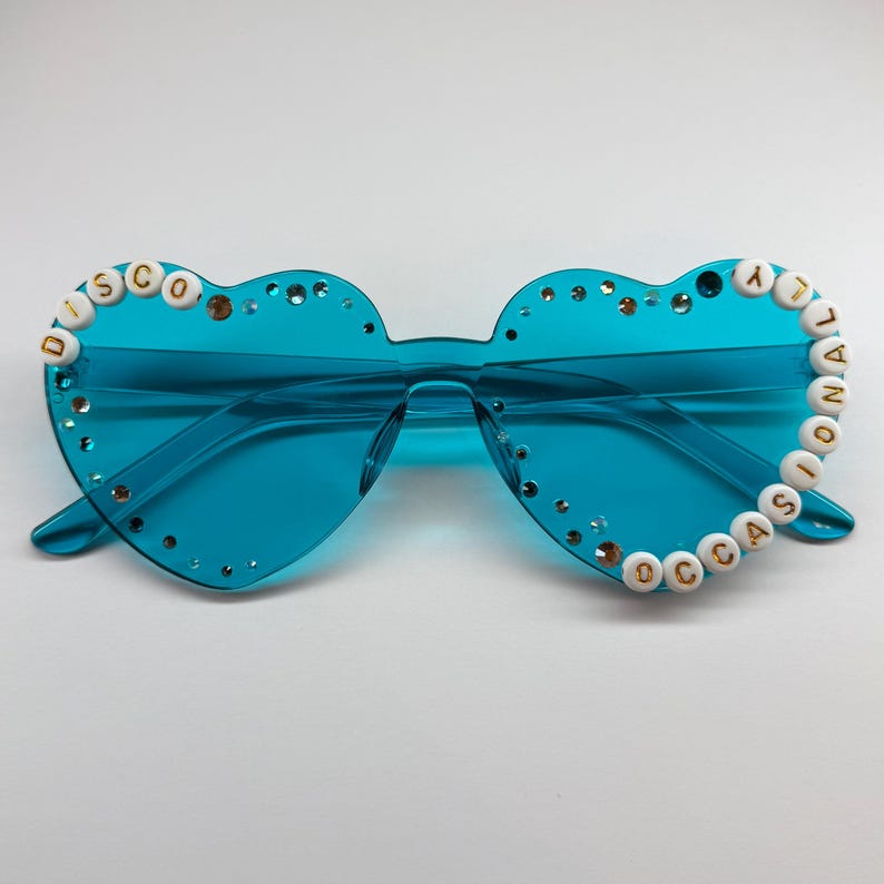 May include: Heart-shaped sunglasses with a vibrant turquoise frame and lenses. The rims are adorned with small, clear rhinestones and the words "DISCO OCCASIONAL" spelled out in white and gold letter beads. A fun, retro accessory.