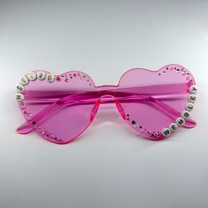 May include: Pink heart-shaped sunglasses with the text "HSTT26" on the left side and "WEMBLEY" on the right side. The sunglasses are adorned with small, sparkling rhinestones around the edges.