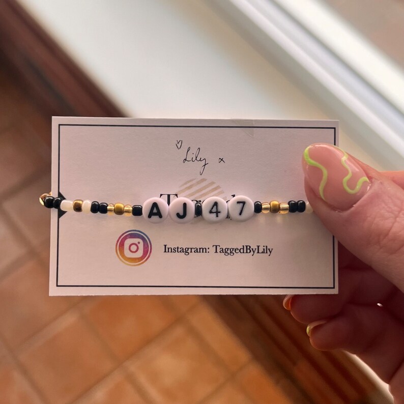 Puede incluir: Una pulsera de cuentas con letras blancas que deletrean "AJ47" se muestra en una tarjeta blanca. La pulsera presenta cuentas negras, doradas y blancas. La tarjeta muestra el nombre de usuario de Instagram "TaggedByLily".