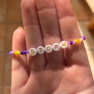Puede incluir: Una pulsera de cuentas con la palabra "STORM" escrita en cuentas de letras blancas con letras doradas. La pulsera presenta una mezcla de cuentas moradas, amarillas y transparentes. La pulsera se sostiene en una mano.