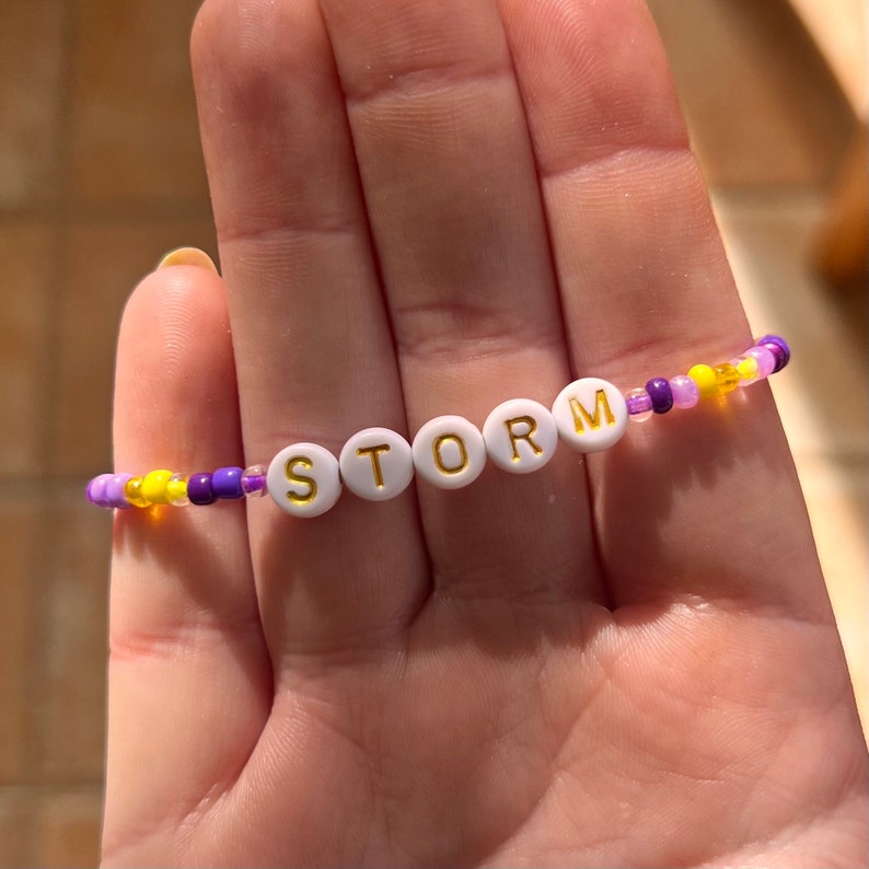 Puede incluir: Una pulsera de cuentas con la palabra "STORM" escrita en cuentas de letras blancas con letras doradas. La pulsera presenta una mezcla de cuentas moradas, amarillas y transparentes. La pulsera se sostiene en una mano.