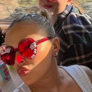 May include: Two people wearing red heart-shaped sunglasses with white embellishments. One person is in the foreground, wearing a gray tank top and a necklace. The other person is in the background, wearing a plaid shirt.