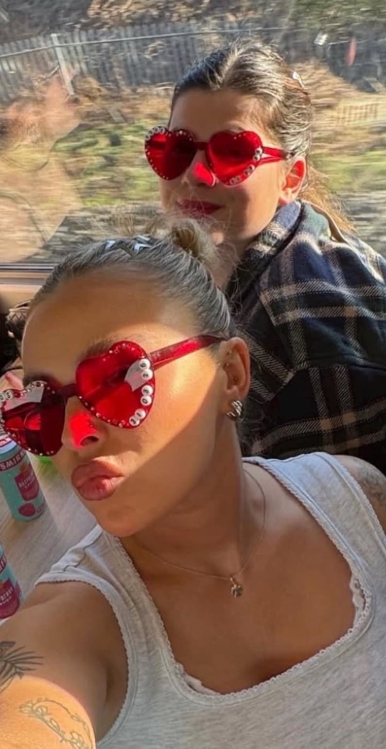 May include: Two people wearing red heart-shaped sunglasses with white embellishments. One person is in the foreground, wearing a gray tank top and a necklace. The other person is in the background, wearing a plaid shirt.