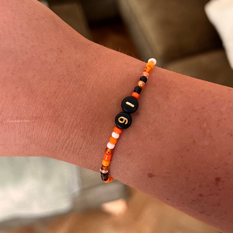 Puede incluir: Una pulsera de cuentas con los n&uacute;meros "19" en dorado sobre cuentas negras. La pulsera presenta un patr&oacute;n de cuentas naranjas, blancas y negras. La pulsera es una joya.