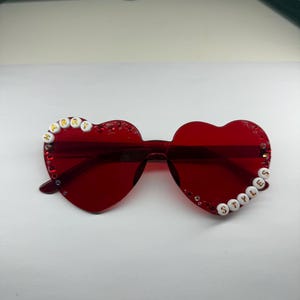 May include: Red heart-shaped sunglasses with the words "HARRY" and "STYLES" spelled out in white beads. The sunglasses have a transparent frame and are decorated with small rhinestones.