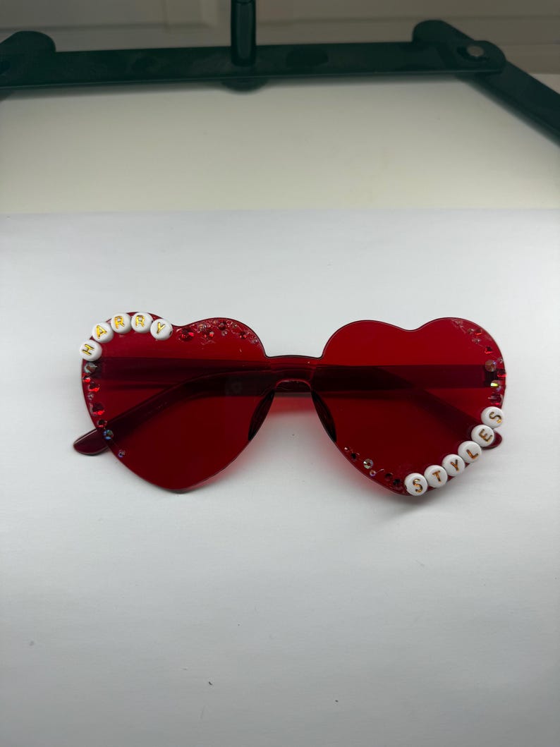 May include: Red heart-shaped sunglasses with the words "HARRY" and "STYLES" spelled out in white beads. The sunglasses have a transparent frame and are decorated with small rhinestones.
