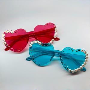 May include: Two pairs of heart-shaped sunglasses. One pair is red with the words "KISS ALL THE TIME" spelled out in white beads. The other pair is blue with the words "DISCO OCCASIONS" in white beads. Both have decorative rhinestones.