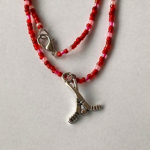 May include: A beaded necklace with a hockey stick charm. The necklace features red and pink beads, with a silver-colored hockey stick pendant. The necklace has a lobster clasp closure.