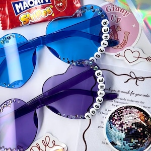 May include: Heart-shaped sunglasses in blue and purple with rhinestone accents. The sunglasses have the word "TOGETHER" spelled out on the frames. The image also includes candy, stickers, and a disco ball.