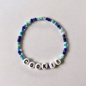 Puede incluir: Una pulsera de cuentas azules y blancas. La pulsera tiene cuentas de letras blancas que deletrean "COOKIE" y el n&uacute;mero "18". Las cuentas est&aacute;n ensartadas en una banda el&aacute;stica.