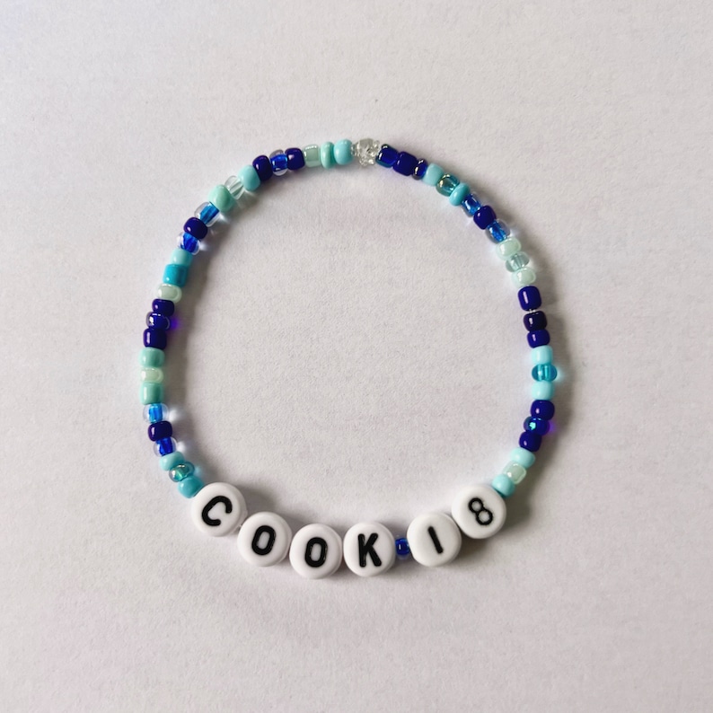 Puede incluir: Una pulsera de cuentas azules y blancas. La pulsera tiene cuentas de letras blancas que deletrean "COOKIE" y el n&uacute;mero "18". Las cuentas est&aacute;n ensartadas en una banda el&aacute;stica.