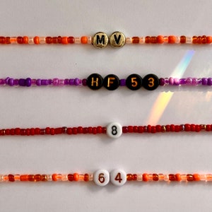 Puede incluir: Cuatro pulseras de cuentas de varios colores. La pulsera superior es naranja y marr&oacute;n con cuentas de letras doradas que dicen "MV". La segunda pulsera es morada con cuentas de letras negras que dicen "HF53". La tercera pulsera es roja con una cuenta blanca que dice "8". La pulsera inferior es naranja con una cuenta blanca que dice "64".