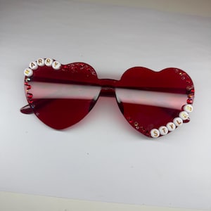 May include: Red heart-shaped sunglasses with the text "HARRY" on the left side and "STYLES" on the right side. The sunglasses have a glittery border and are a vibrant shade of red.