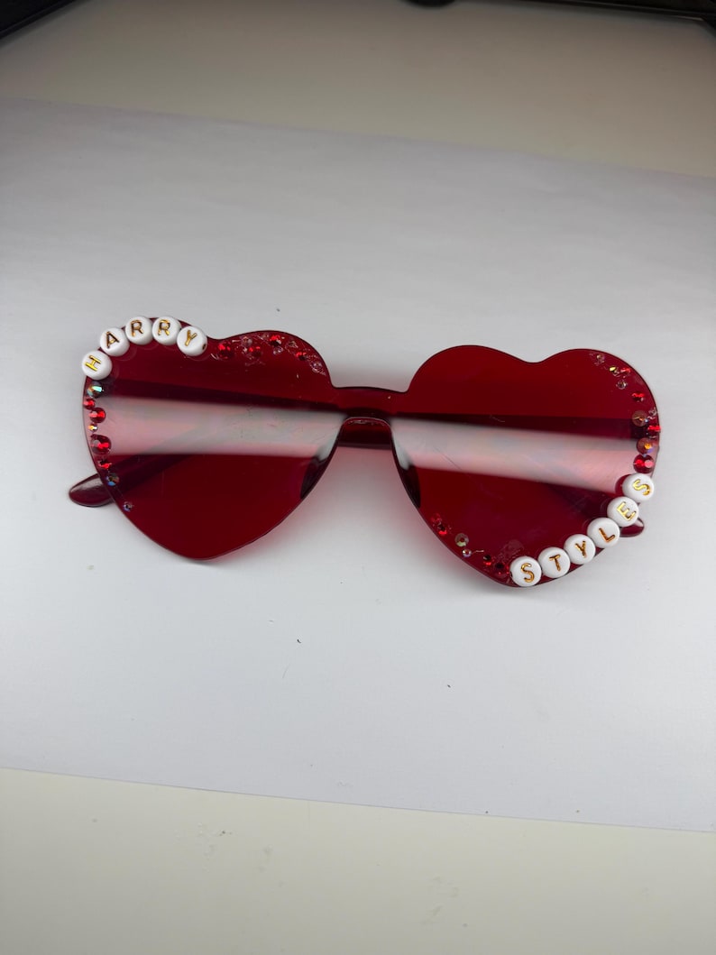 May include: Red heart-shaped sunglasses with the text "HARRY" on the left side and "STYLES" on the right side. The sunglasses have a glittery border and are a vibrant shade of red.