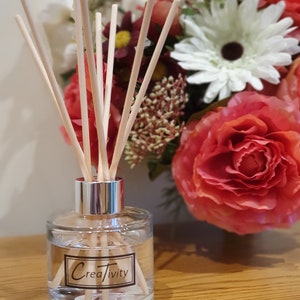 May include: A clear glass bottle diffuser with a silver cap and wooden sticks. The bottle has a black label with the word "Creativity" on it. The diffuser is sitting on a wooden surface in front of a bouquet of pink and white flowers.