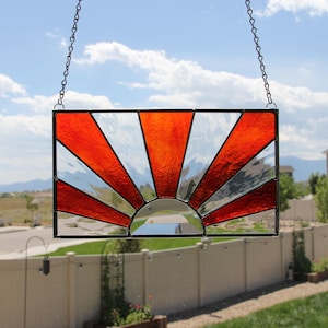May include: Rectangular stained glass panel featuring a rising sun design. The sun's rays are depicted in vibrant red and clear glass, radiating from a semi-circular base. The panel is framed in black and suspended by chains.
