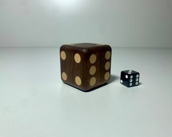 May include: Two dice, one made of wood with gold dots and one black with white dots. The wooden die is larger than the black die.