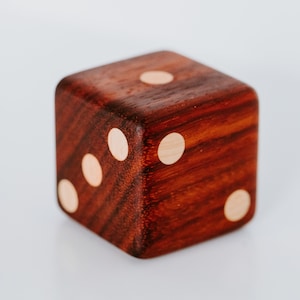 May include: A wooden dice with three white dots on one side and two white dots on another side. The dice is made of dark wood with a reddish hue.