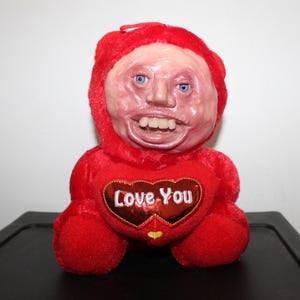 May include: A red plush toy with a human-like face and a heart-shaped patch that says "Love You".