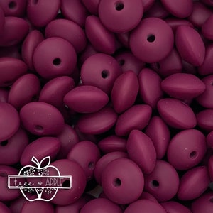 May include: A close-up shot of a pile of plum-colored beads. The beads are a mix of round and oval shapes, each with a hole through the center. The image includes the text "tree & APPLE" in a white logo.