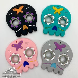 May include: Four colorful silicone sugar skull teethers. The teethers are black, teal, pink, and gray with white, purple, and orange accents. Each teether has two holes for fingers.